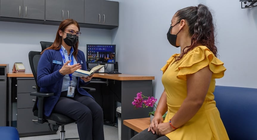 Una consejera espiritual de la Clínica Bíblica con mascarilla conversa y guía a una paciente usando una Biblia en un consultorio médico.