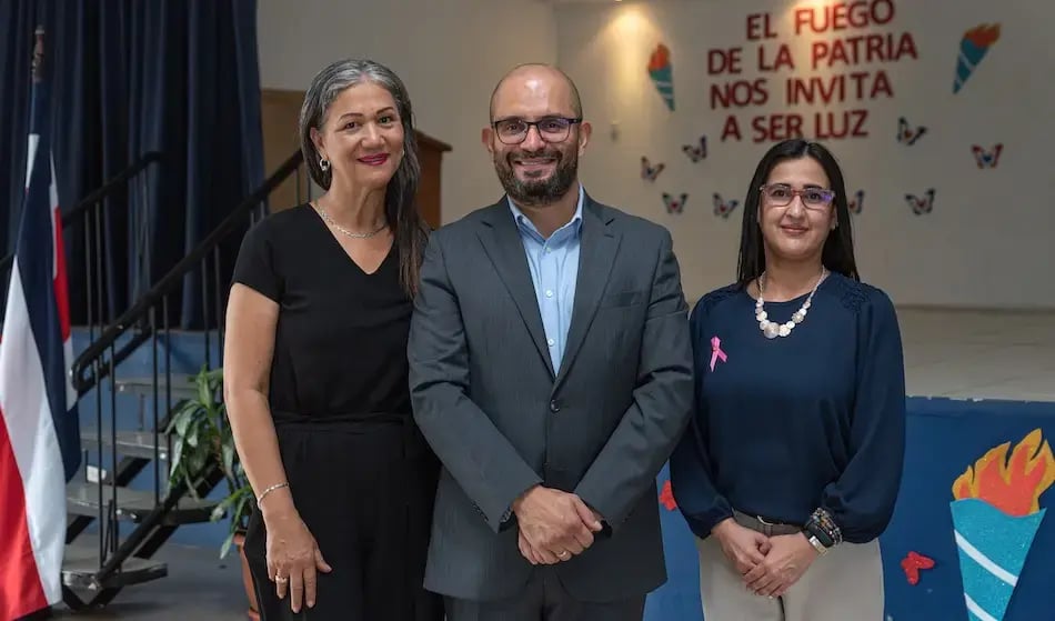Tres adultos posando frente a una tarima decorada con motivos patrios en una escuela.