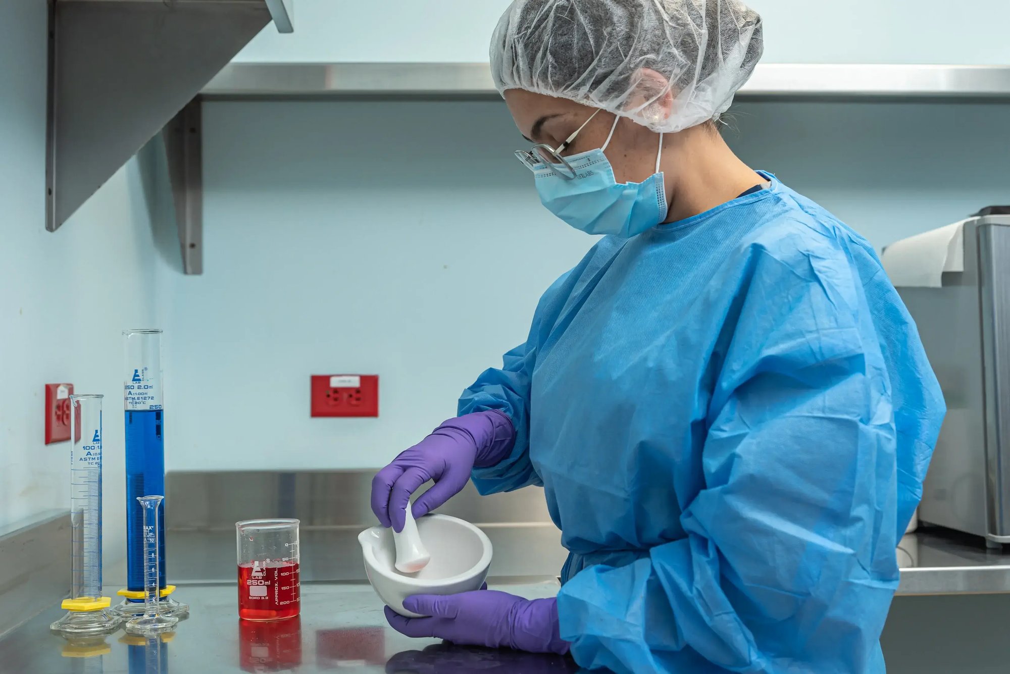 Analista de laboratorio preparando muestra en mortero junto a vasos de precipitados con líquidos de colores.
