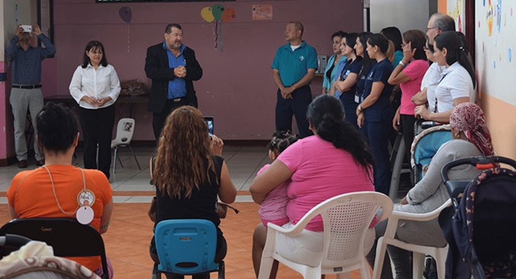 Reunión comunitaria con personas sentadas y de pie durante una actividad en un salón interior.
