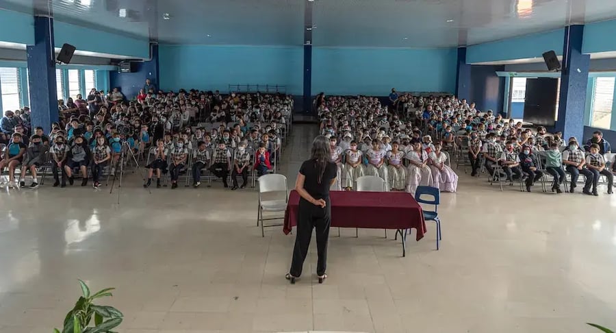 Niños y niñas sentados en un auditorio escolar mientras una mujer de pie les habla desde el frente.