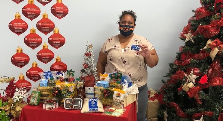 Una mujer con mascarilla sostiene una tarjeta de regalo junto a una mesa llena de productos alimenticios básicos y un árbol de Navidad, como parte de la campaña Ilumina una mesa.