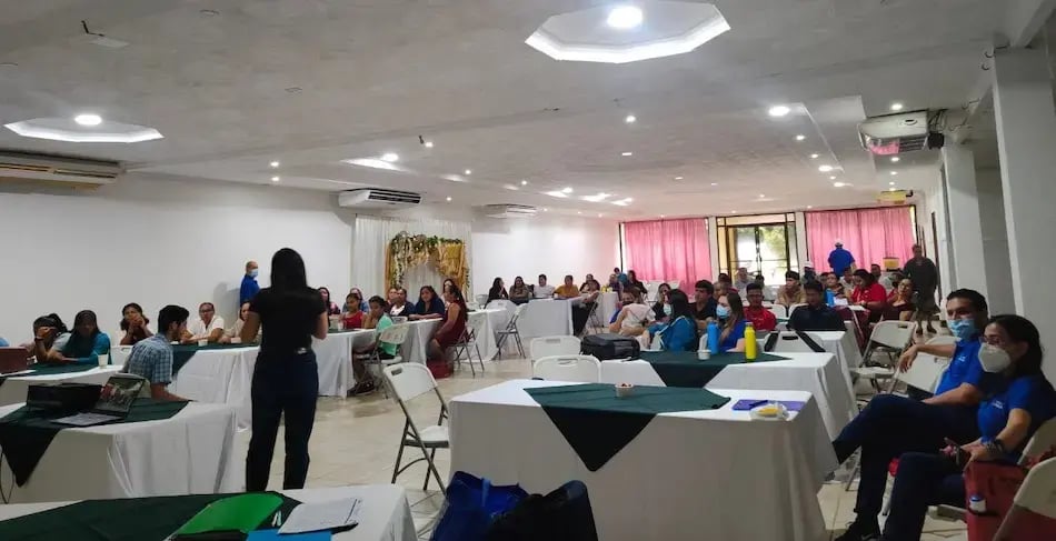 Grupo numeroso de personas sentadas en mesas mientras escuchan una charla en un salón amplio.