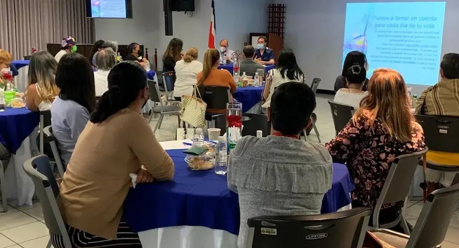 Personas participando en una charla grupal en un salón, sentadas alrededor de mesas mientras escuchan una presentación proyectada.