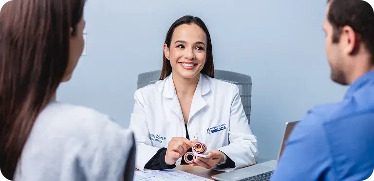 Una médica especialista de Clínica Bíblica le muestra y explica un modelo anatómico a una pareja sentada en el consultorio.