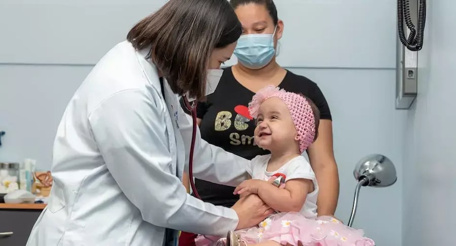 Médica examinando a una bebé sentada en la camilla mientras la madre observa en el consultorio.