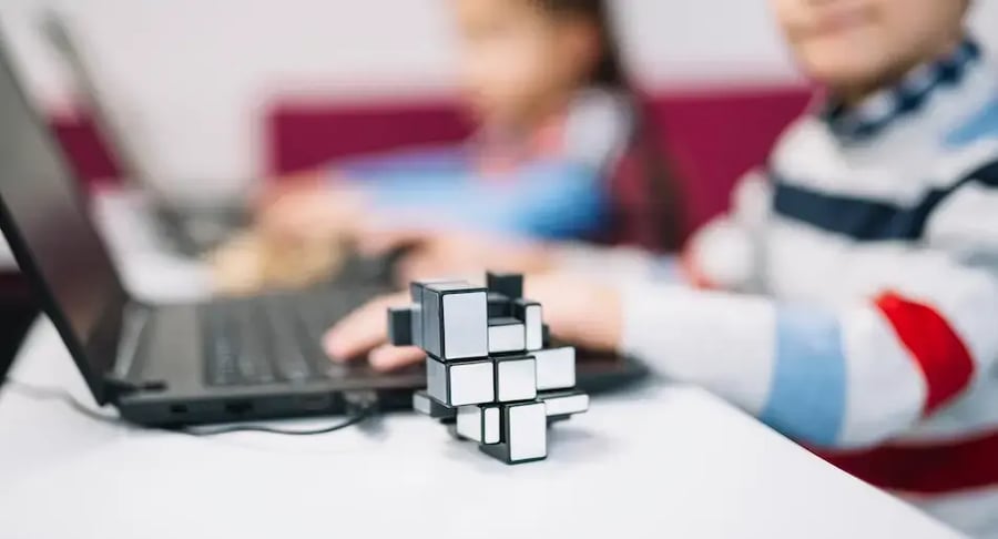 Cubo metálico de rompecabezas sobre una mesa junto a una laptop, con niños usando la computadora al fondo desenfocado.