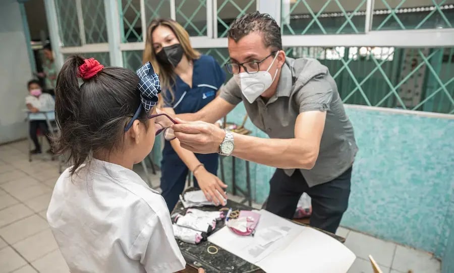 Hombre colocando lentes a una niña en un aula escolar mientras una profesional observa.