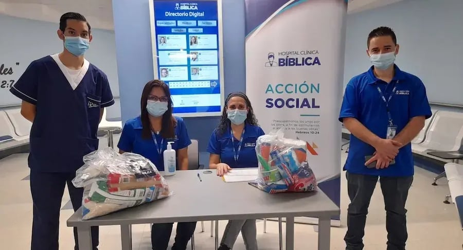 Equipo de Acción Social de la Clínica Bíblica sentado y de pie junto a una mesa con paquetes de víveres en un pasillo del hospital.
