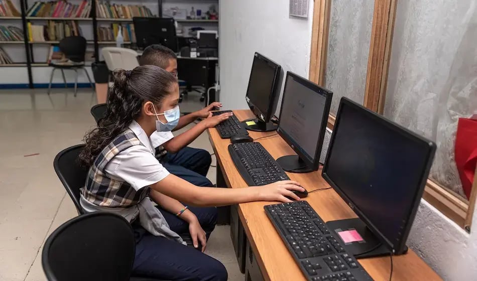 Estudiantes con uniforme escolar usando computadoras de escritorio en un laboratorio de cómputo.