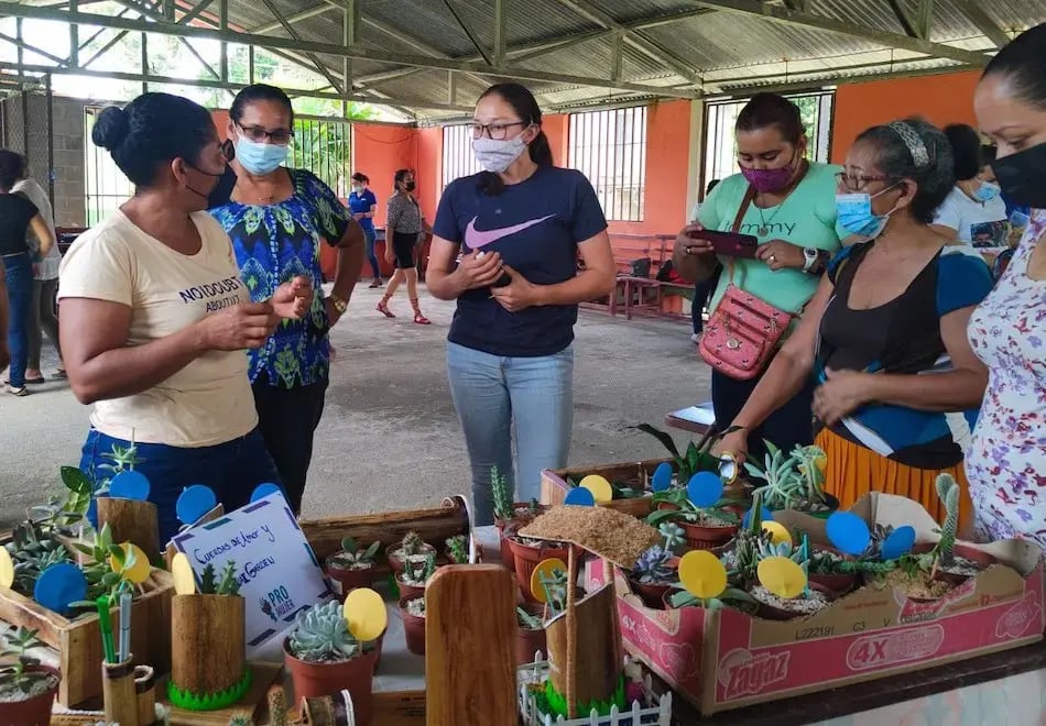 Varias mujeres observando y conversando sobre plantas decorativas en macetas durante una feria comunitaria.