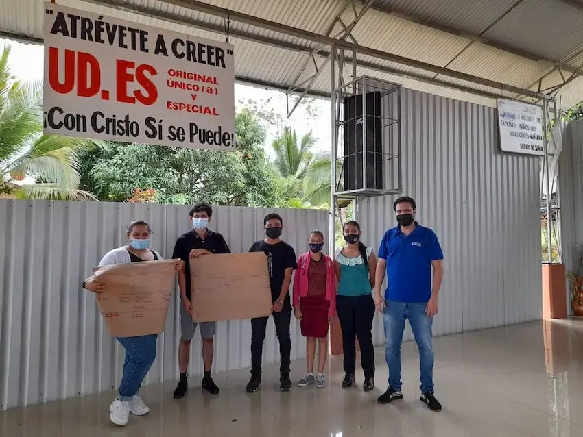 Grupo de jóvenes y un instructor posan de pie bajo un rótulo, sosteniendo carteles grandes de papel en un salón techado.