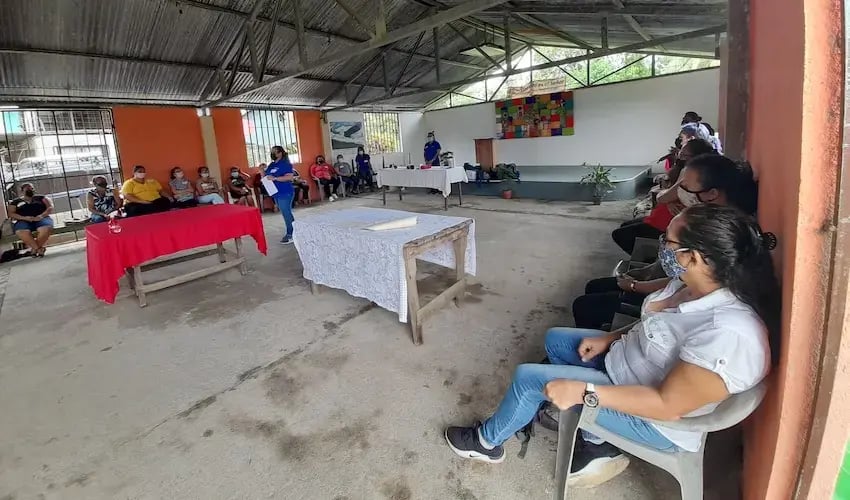 Mujer con camisa azul dando una charla a un grupo de mujeres sentadas en sillas dentro de un salón techado.