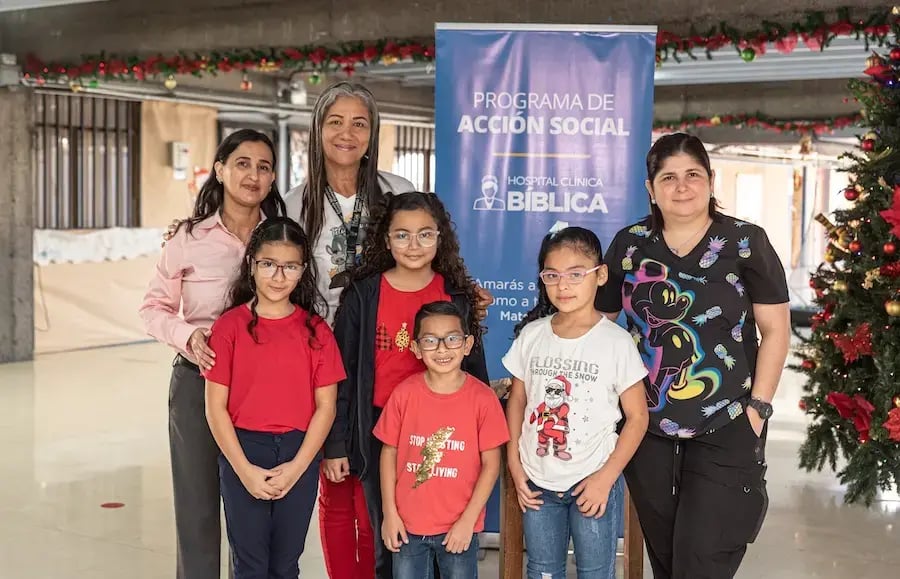 Grupo de mujeres y niños posando frente a un rótulo del Programa de Acción Social junto a un árbol decorado.