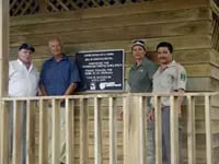Cuatro personas posando frente a una estructura de madera con una placa conmemorativa