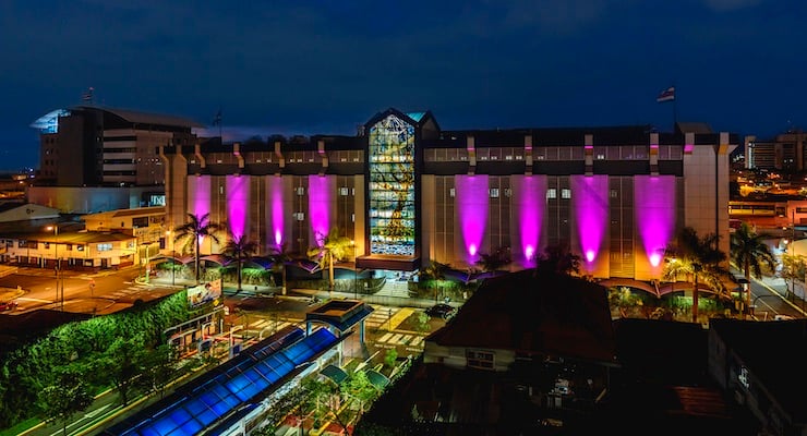 Vista nocturna de la fachada principal del Hospital Clínica Bíblica iluminada con luces color rosa en conmemoración del mes de la lucha contra el cáncer de mama.