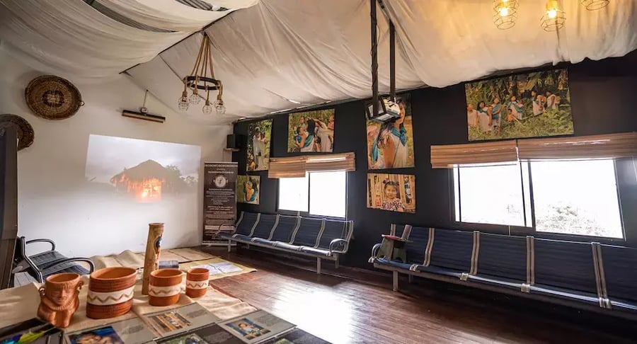 Sala de exhibición con bancas, proyección en la pared, piezas artesanales y fotografías de actividades culturales.