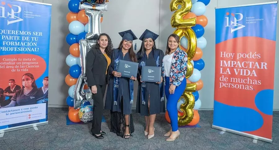 Dos graduadas con toga y birrete posando con sus diplomas junto a dos acompañantes frente a decoraciones y paneles informativos del Instituto Panamericano AsemeCo.
