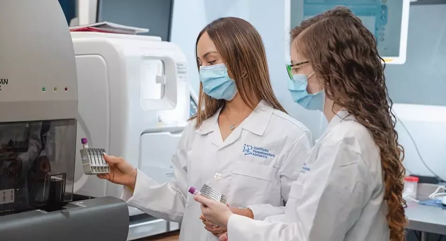 Dos profesionales de laboratorio con bata y mascarilla revisando tubos de muestra frente a un equipo analizador.