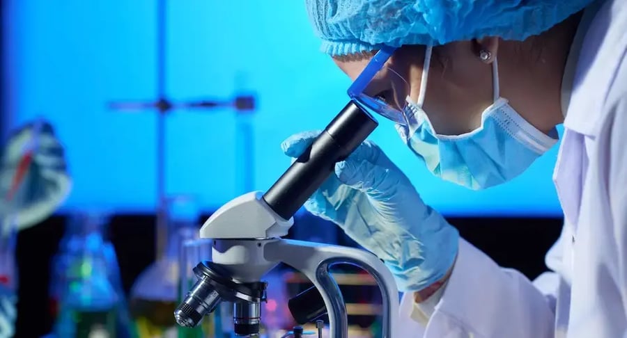 Investigadora con mascarilla y guantes observando muestras a través de un microscopio en un laboratorio.