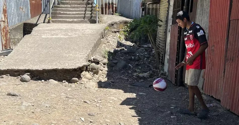 Joven dominando un balón de fútbol en un área de tierra frente a una vivienda con paredes de zinc.