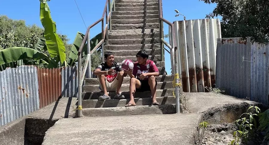 Dos jóvenes sentados en unas gradas de cemento conversando mientras sostienen un balón de fútbol.