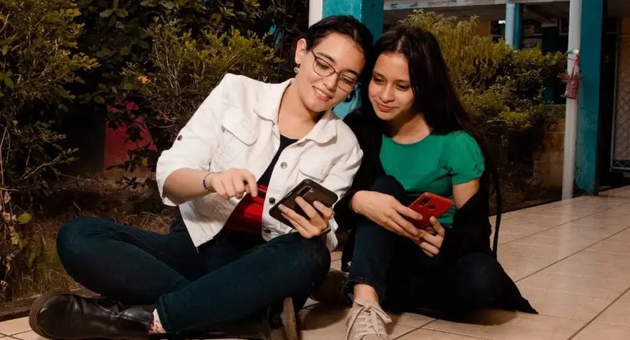 Dos jóvenes sentadas en el suelo usando sus teléfonos móviles mientras comparten y observan la pantalla juntas.