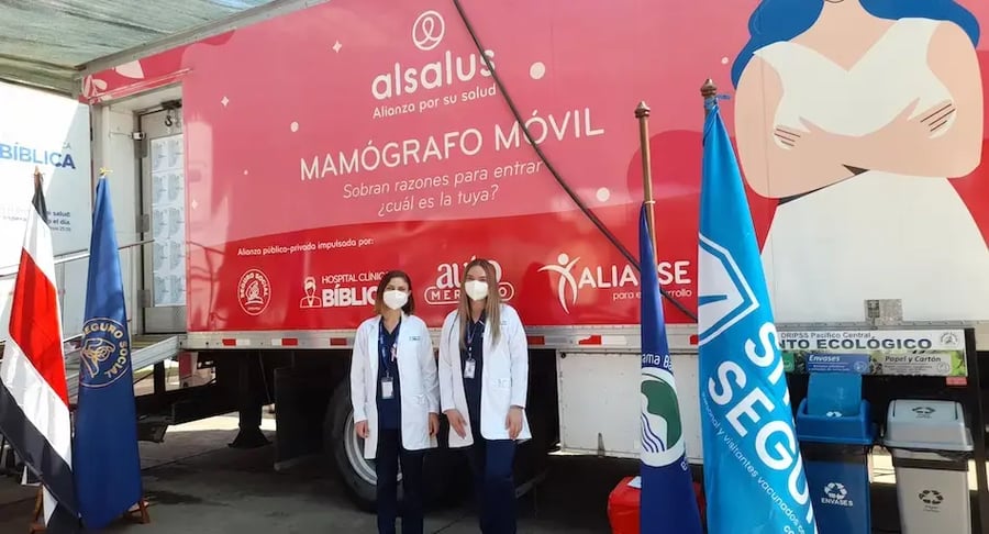 Dos profesionales de salud con batas blancas posan frente a un mamógrafo móvil junto a banderas institucionales.