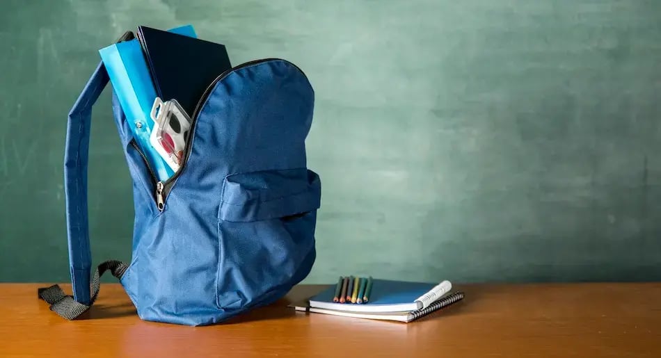 Mochila escolar azul abierta con útiles y cuadernos sobre un escritorio frente a un pizarón.
