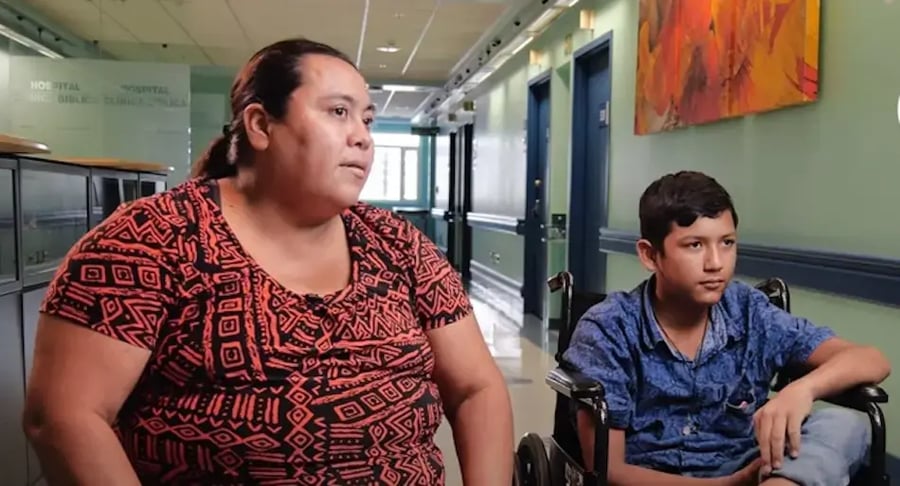 Mujer sentada junto a un adolescente en silla de ruedas en un pasillo de hospital.