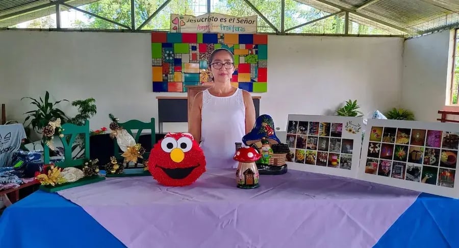Mujer junto a una mesa exhibiendo artesanías y figuras decorativas en un salón techado.