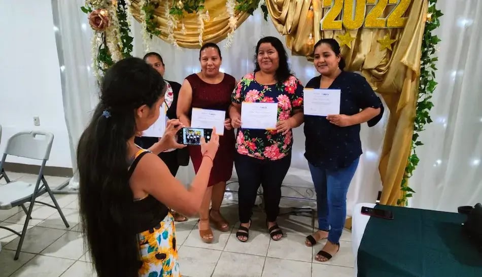Tres mujeres posando con certificados mientras otra persona toma una foto con su teléfono.