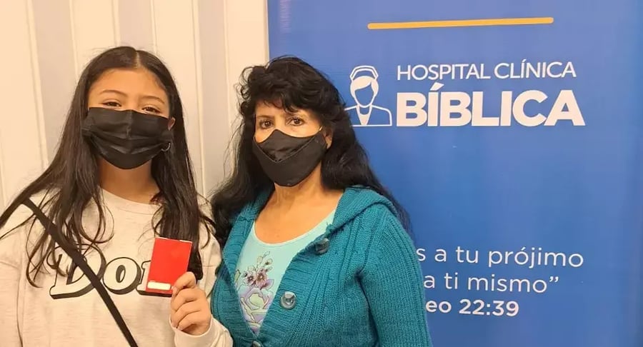 Dos mujeres con mascarilla posando frente a un rótulo de Hospital Clínica Bíblica mientras una de ellas sostiene una tarjeta roja.