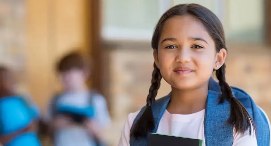 Niña con mochila azul sonriendo mientras sostiene un cuaderno en un entorno escolar.