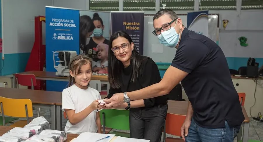 Niña recibiendo un obsequio de dos adultos en un aula con rótulos de programas sociales en el fondo.