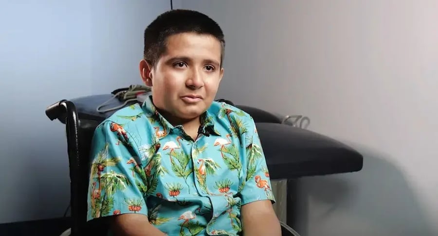 Niño sentado en una silla de ruedas dentro de un consultorio médico, usando una camisa estampada.