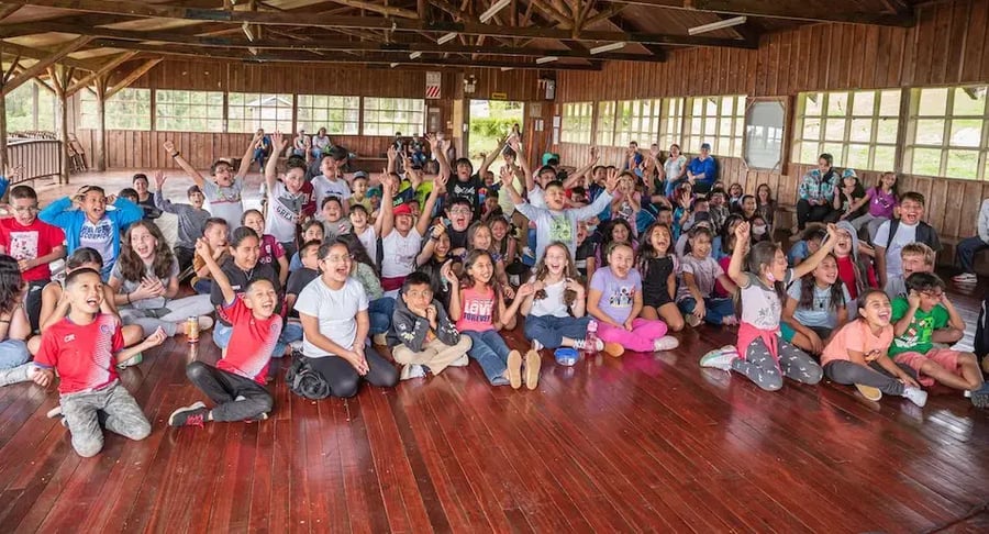 Grupo numeroso de niños sentados en un salón de madera celebrando y levantando los brazos.