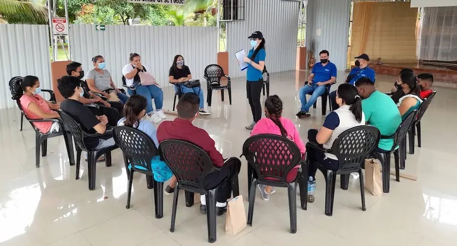 Niños y jóvenes sentados en círculo en un salón amplio mientras una facilitadora con mascarilla habla frente al grupo.