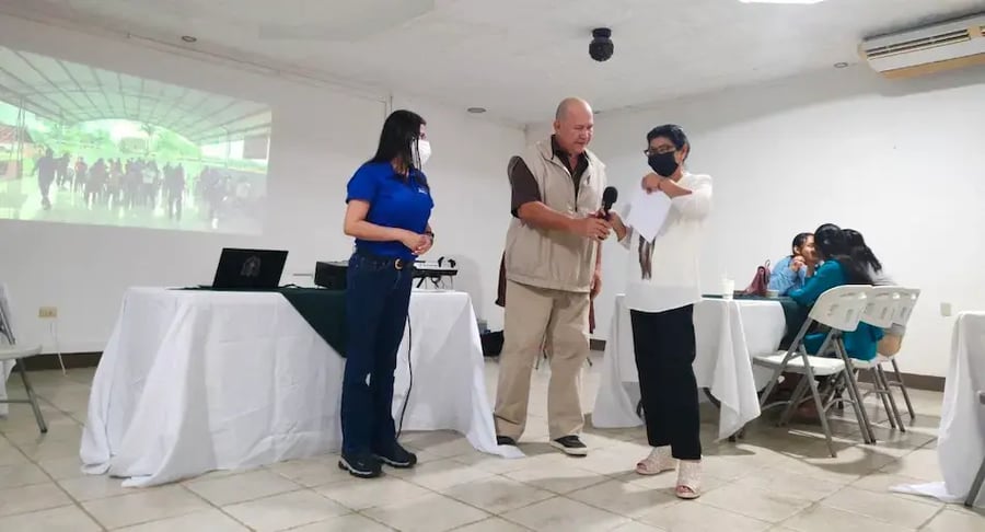 Tres personas de pie frente a un grupo durante una presentación en un salón con proyector.