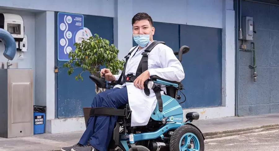 Profesional de salud con bata y mascarilla usando una silla de ruedas eléctrica en el exterior de un edificio.