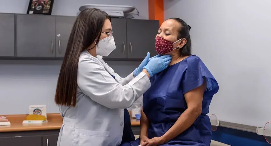 Doctora con mascarilla y guantes examinando el cuello de una paciente en una sala de consulta.