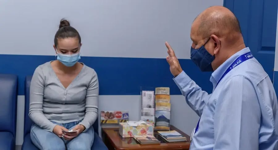 Mujer con mascarilla sentada en sala de espera mientras un funcionario con mascarilla le dirige un saludo.