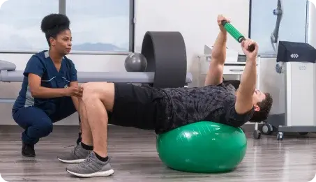 Un paciente realiza ejercicios de rehabilitación sobre una bola Suiza mientras una terapeuta física supervisa su postura.
