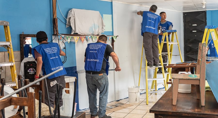 Voluntarios con chalecos azules realizando labores de pintura y reparación en el interior de un salón con escaleras y herramientas.