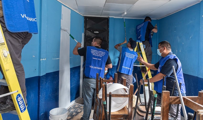 Grupo de voluntarios pintando paredes y realizando arreglos dentro de un salón comunitario con muebles apilados.