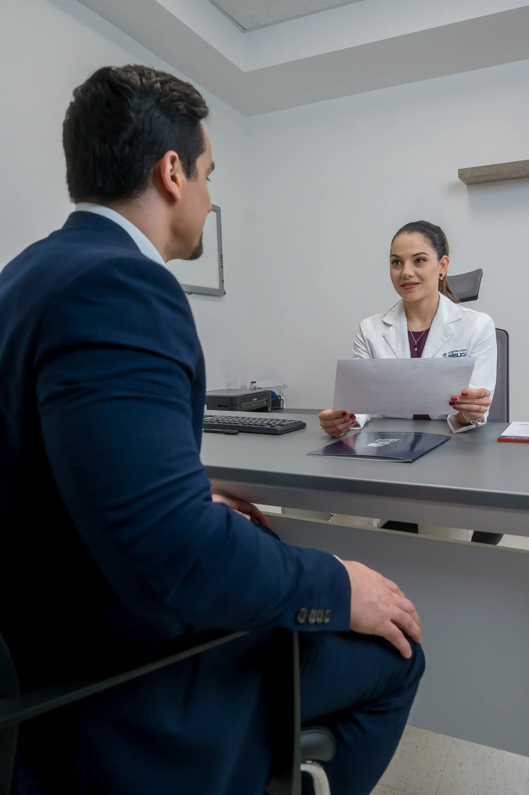 Médica mostrando un documento a un paciente sentado frente al escritorio en un consultorio.