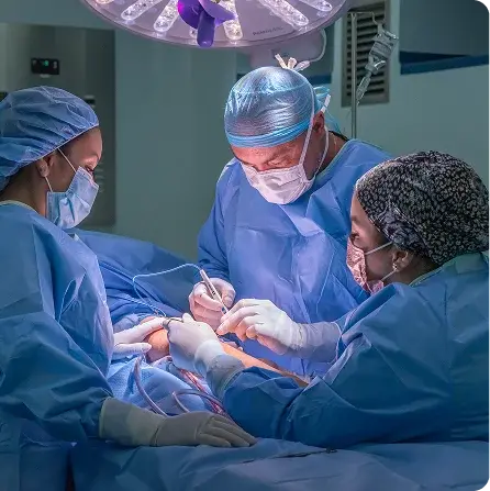 Tres profesionales de salud (dos médicas y un médico cirujano) con uniformes quirúrgicos azules, gorros y mascarillas, operan a un paciente bajo una lámpara de quirófano, enfocados en un procedimiento delicado.