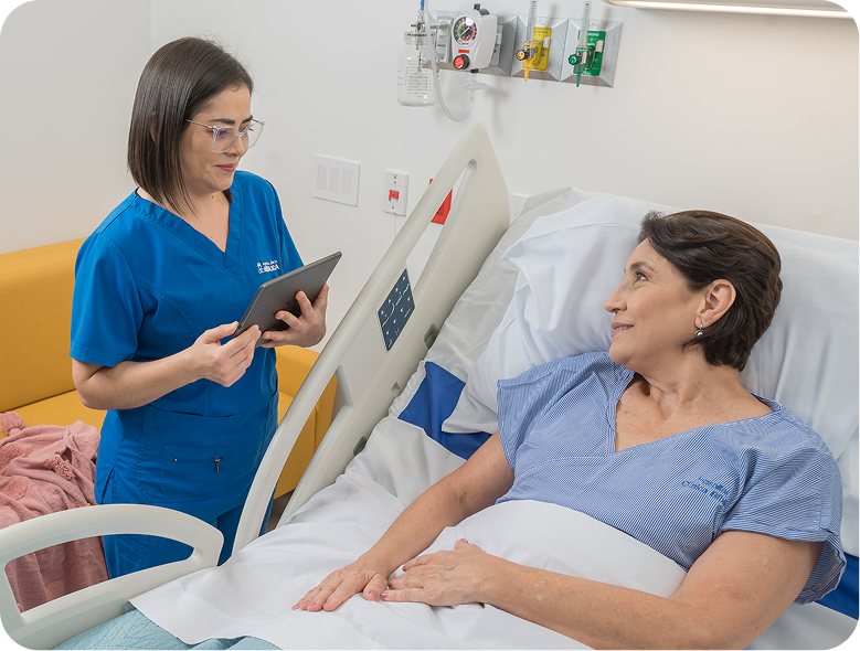 Enfermera con uniforme azul sostiene una tableta mientras conversa con una paciente recostada en una cama de hospital.