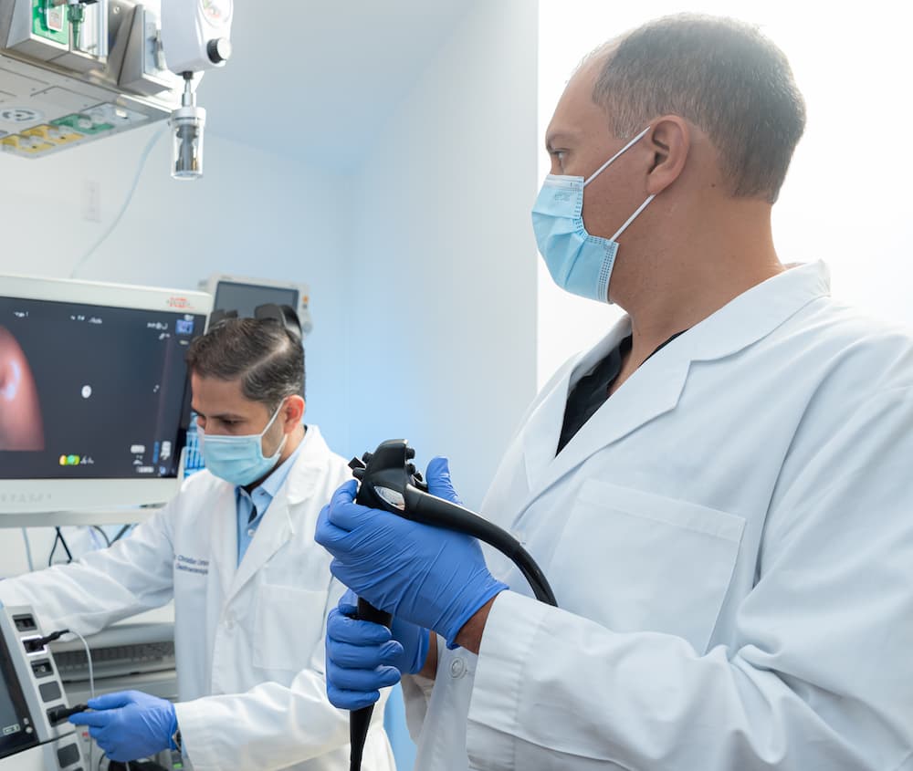 Foto de medicos gastroenterologos realizando un procedimiento de endoscopia. Un doctor sostiene el endoscopio con guantes y mascarilla de proteccion, mientras su colega opera el equipo medico frente a una pantalla de monitoreo.