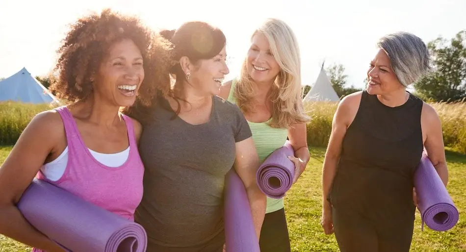Group of smiling adult women with yoga mats initiating outdoor physical activity.